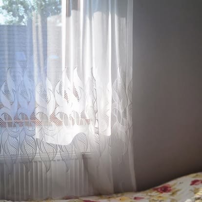 Firana i grzejnik przy oknie w pokoju, w rogu narzuta na łóżku